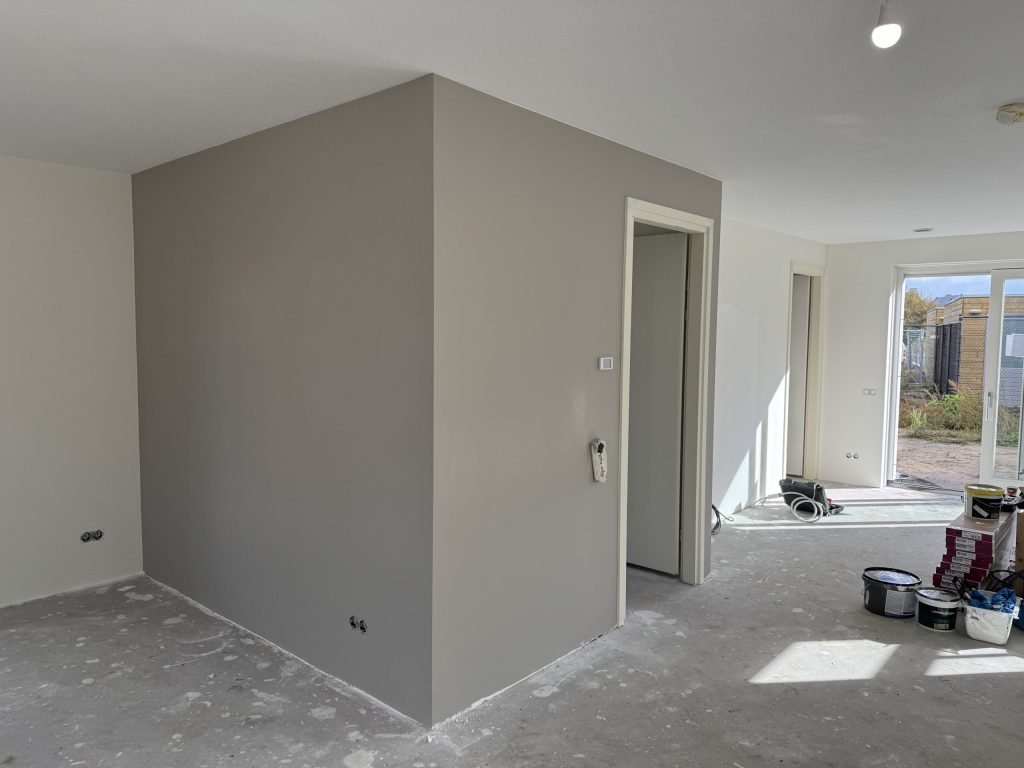 Woonkamer met een net gesausde grijze wand rond de meterkast, verfemmers nog op de vloer